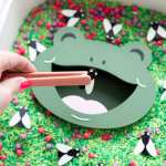 A close-up of a sensory bin filled with green dyed rice, red and purple loose parts, and black and white bug figures. A green frog face insert with an open mouth sits in the center. A hand with bright pink nails holds peach tweezers, placing a bug piece into the frog's mouth.