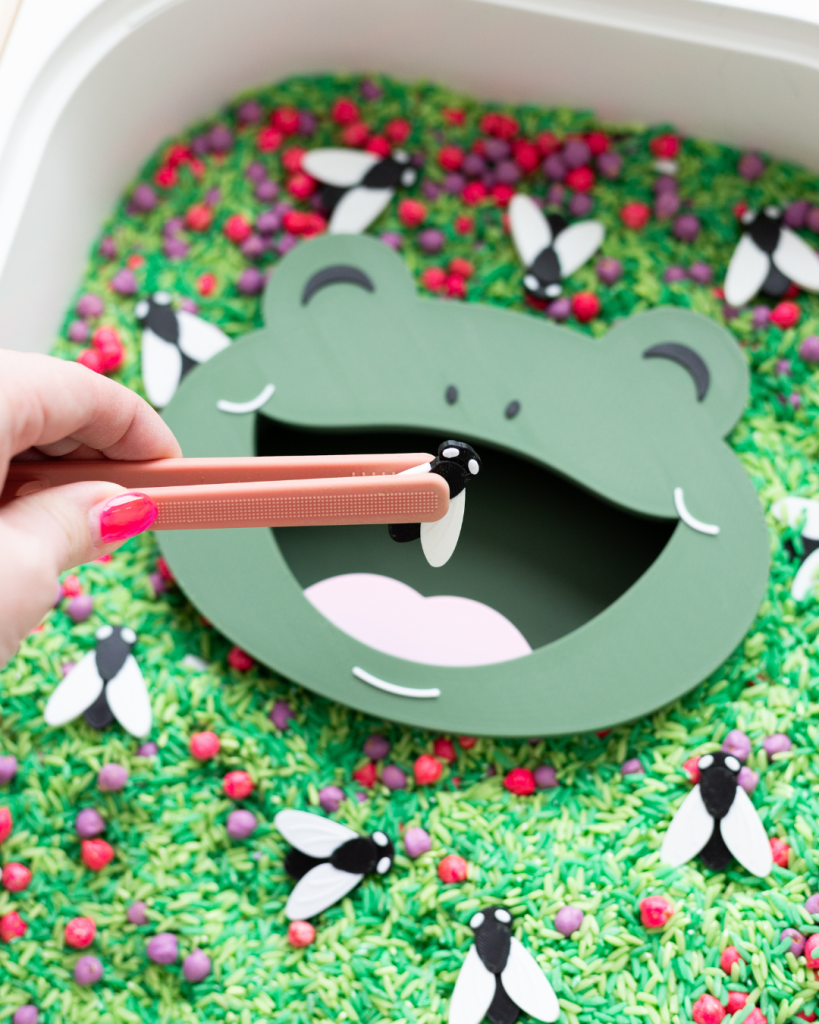 A close-up of a sensory bin filled with green dyed rice, red and purple loose parts, and black and white bug figures. A green frog face insert with an open mouth sits in the center. A hand with bright pink nails holds peach tweezers, placing a bug piece into the frog's mouth.