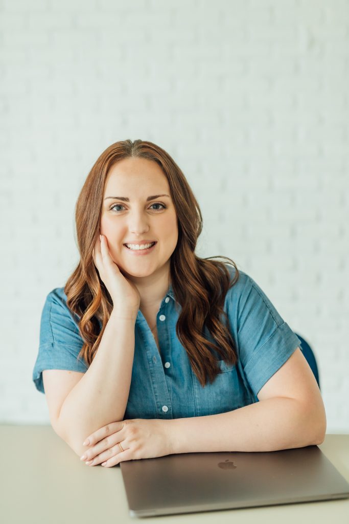 Ashlee Wiggins, maker and creator behind by.ashwiggins, smiling at a desk with a MacBook against a white brick background