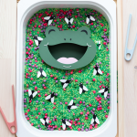 A close-up of a sensory bin filled with green dyed rice, red and purple loose parts, and black and white bug figures. A green frog face insert with an open mouth sits in the center. A hand with bright pink nails holds peach tweezers, placing a bug piece into the frog's mouth.