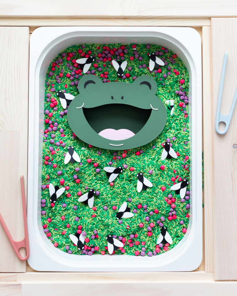 A close-up of a sensory bin filled with green dyed rice, red and purple loose parts, and black and white bug figures. A green frog face insert with an open mouth sits in the center. A hand with bright pink nails holds peach tweezers, placing a bug piece into the frog's mouth.