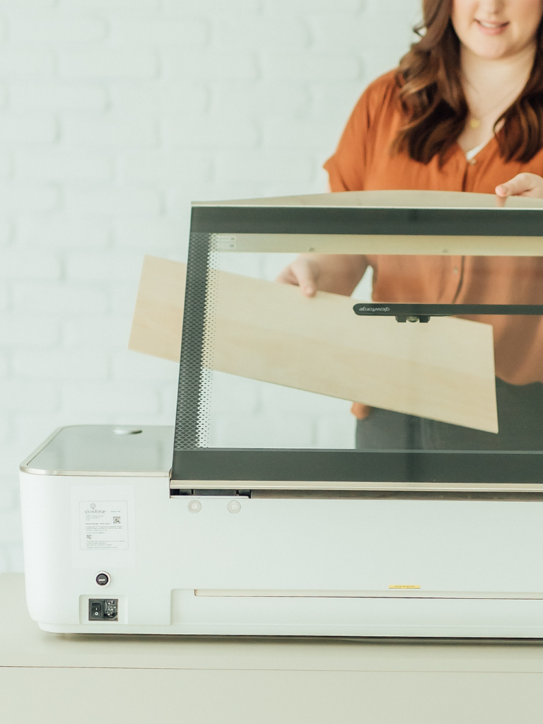A woman with brown hair places a rectangular sheet of light wood into the open lid of a white Glowforge laser cutter, set against a white brick wall background.