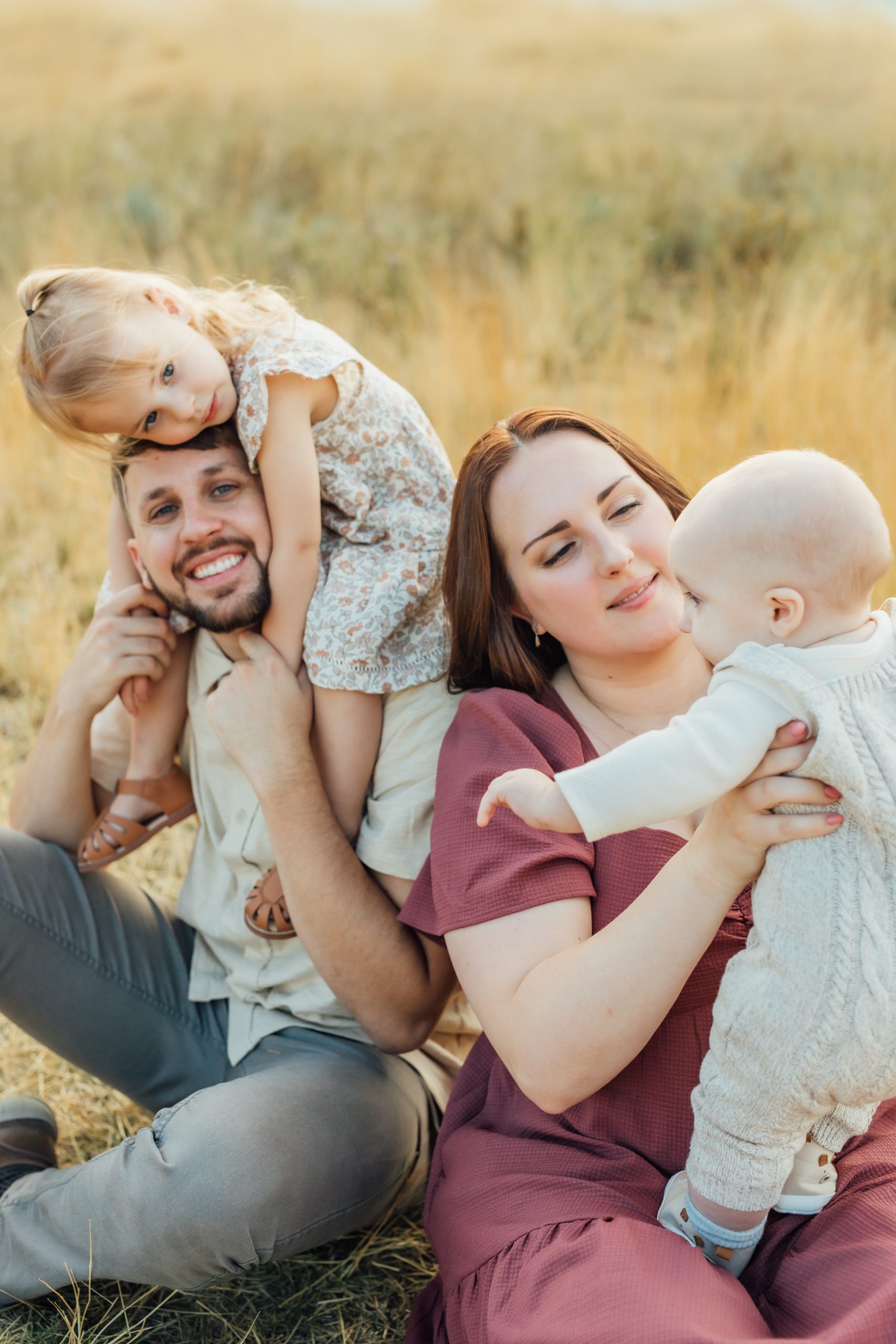 A mom and dad sit in a sunny field with their toddler daughter climbing on dad's back and their baby leaning in for a kiss.