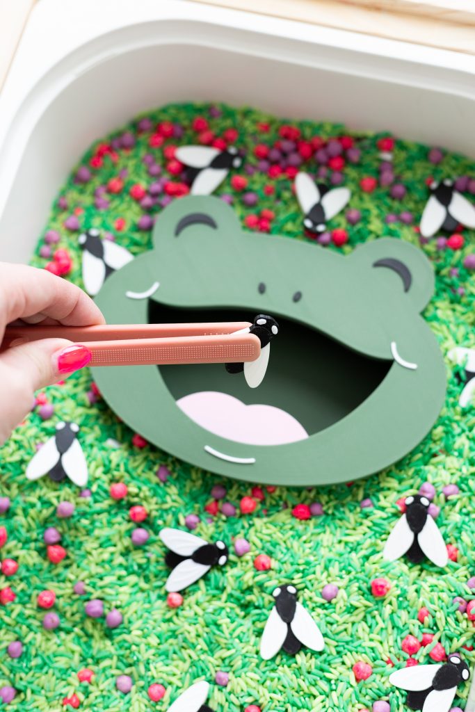 A close-up of a sensory bin filled with green dyed rice, red and purple loose parts, and black and white bug figures. A green frog face insert with an open mouth sits in the center. A hand with bright pink nails holds peach tweezers, placing a bug piece into the frog's mouth.
