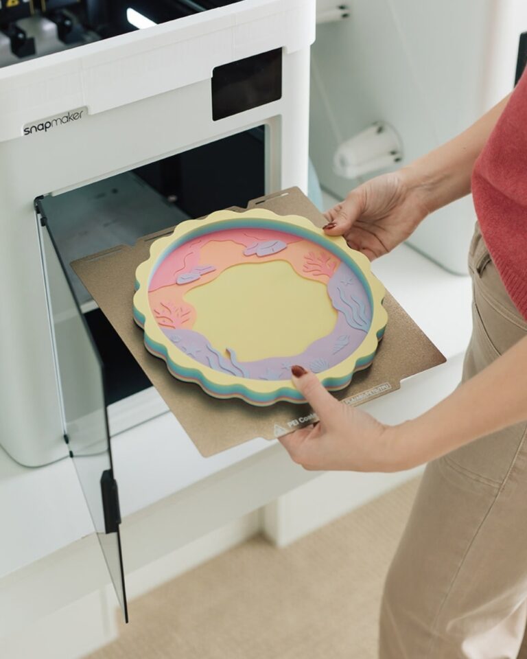 Ash holding a finished 3D printed Mermaid Treasure Trove Tray with pastel rainbow layers in front of a 3D printer.